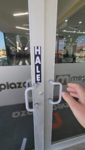 A person opening a glass door with a white handle at a business entrance in Roatan, Honduras, with a vertical "SALE" sign attached to the door frame.