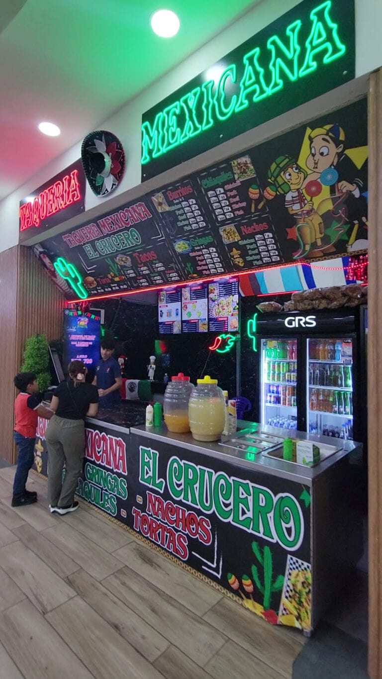 A colorful Mexican food stand in Roatan, Honduras, features neon signs, a menu with tacos, nachos, and tortas, and customers ordering while a staff member prepares food behind the counter.