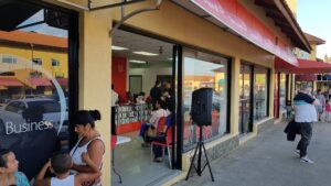 People gather outside a storefront with large glass windows on a busy street in Roatan, Honduras, showing a mix of customers sitting inside and pedestrians walking by on a sunny day.