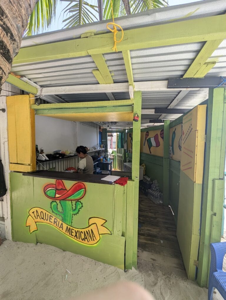 A colorful Mexican taquería named "Taquería Mexicana" features a green wooden booth with a yellow banner, situated outdoors on a sandy surface in Roatan, Honduras, with palm leaves visible overhead.