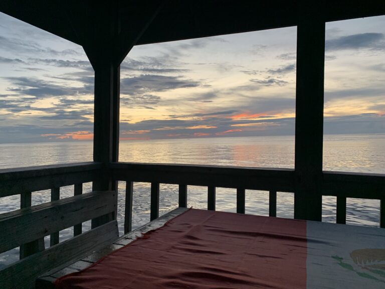 A wooden deck with seating overlooks the calm waters of Roatan, Honduras during sunset, with a partly cloudy sky and colorful horizon in the background.