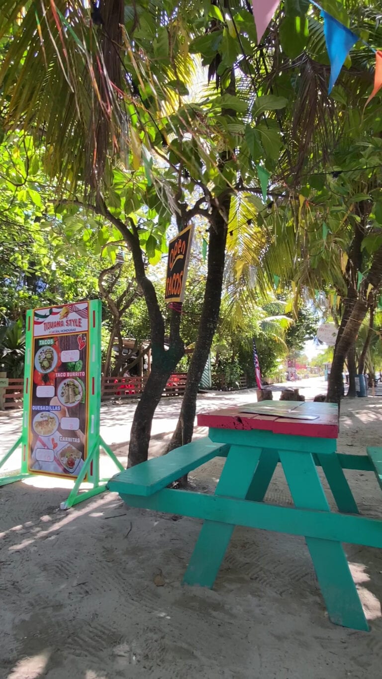 A colorful outdoor dining area on a sandy beach in Roatan, Honduras, featuring a turquoise picnic table, a menu sign advertising tacos, burritos, quesadillas, and carnitas, surrounded by lush tropical trees and banners hanging from the foliage.