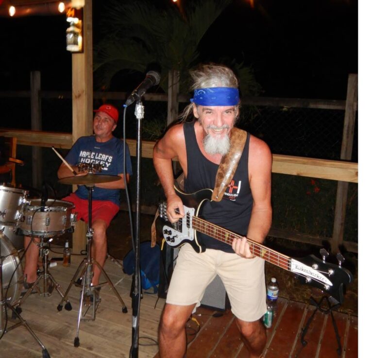 A lively musician with a gray beard and blue headband plays an electric guitar during a nighttime performance at an outdoor venue in Roatan, Honduras, while a drummer wearing a red cap and "Maple Leafs Hockey" shirt accompanies him.