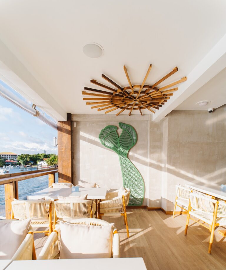 A shaded outdoor seating area on Roatan, Honduras, features white wicker chairs, a green mermaid wall sculpture, and a wooden sunburst ceiling decor with a view of boats and water in the background.