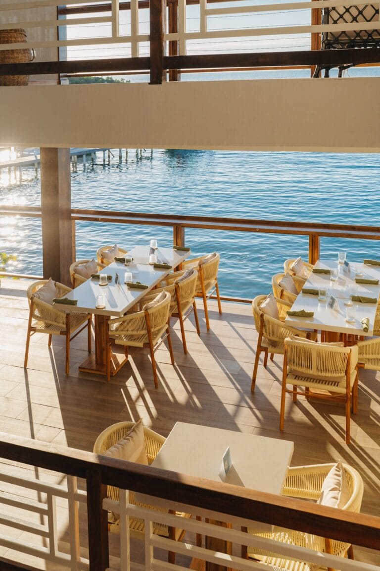 A seaside restaurant on Roatan, Honduras, features outdoor dining with white tables, beige wicker chairs with cushions, and a view of the ocean through wooden and white railing balconies.