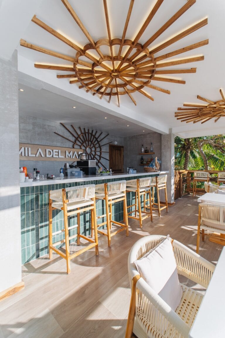 A modern indoor bar area on Roatan, Honduras features a wooden sunburst ceiling light fixture, a bar with green tiled front, white bar stools, and greenery visible through an open balcony space.