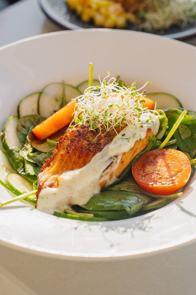 A plated dish featuring a grilled fillet of fish topped with creamy white sauce, garnished with microgreens, served with slices of cucumber, cherry tomatoes, and roasted vegetables, on a white ceramic plate in Roatan, Honduras.