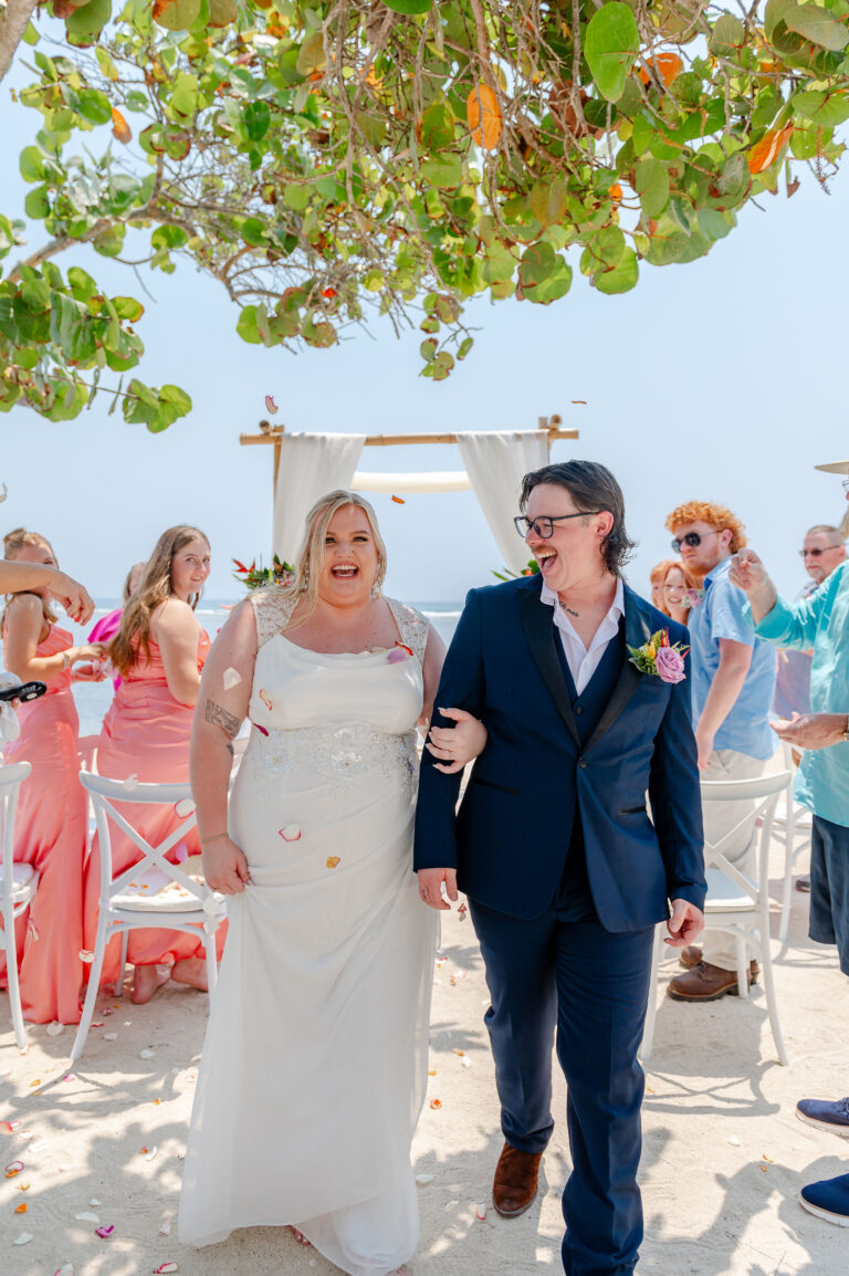 A newlywed couple walking hand in hand on the sandy beach in Roatan, Honduras during their wedding celebration with friends and family around under the shade of a leafy tree.