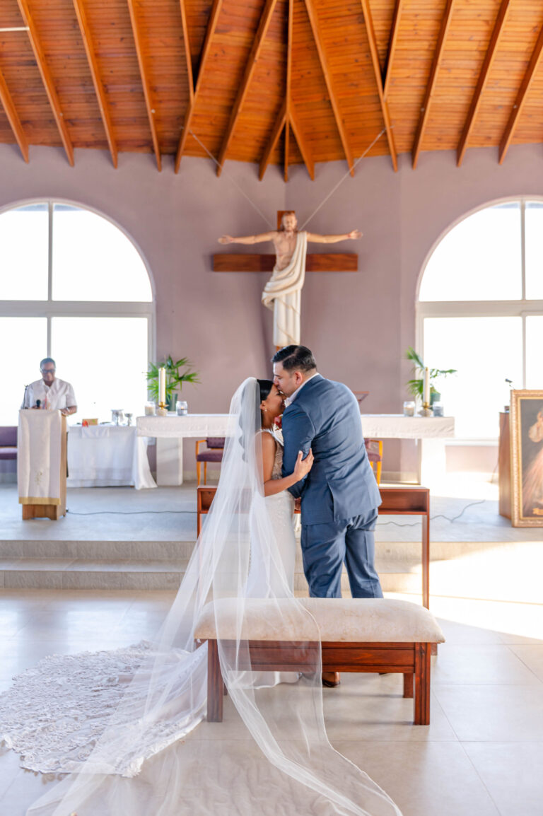 A bride and groom share a kiss during their wedding ceremony inside a church in Roatan, Honduras, with a prominent crucifix and large arched windows behind them.