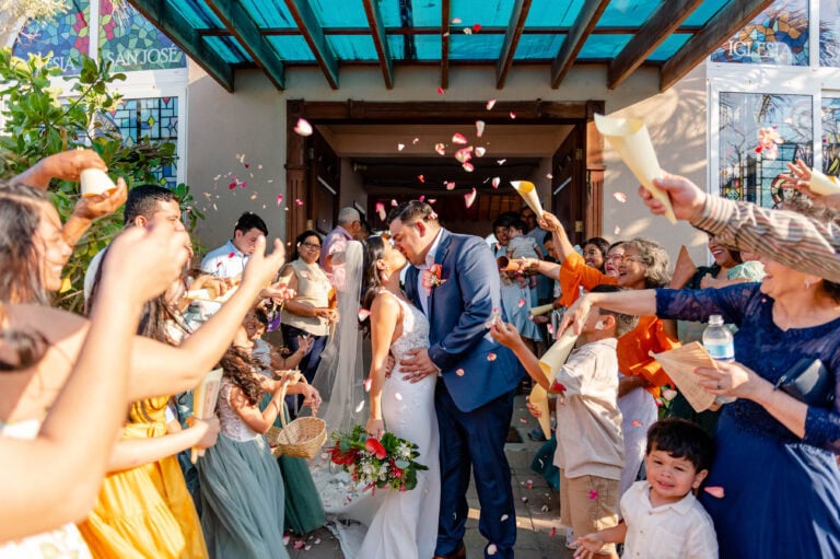 A newlywed couple shares a kiss in front of a crowd of cheerful guests tossing flower petals during a wedding celebration at a church in Roatan, Honduras.