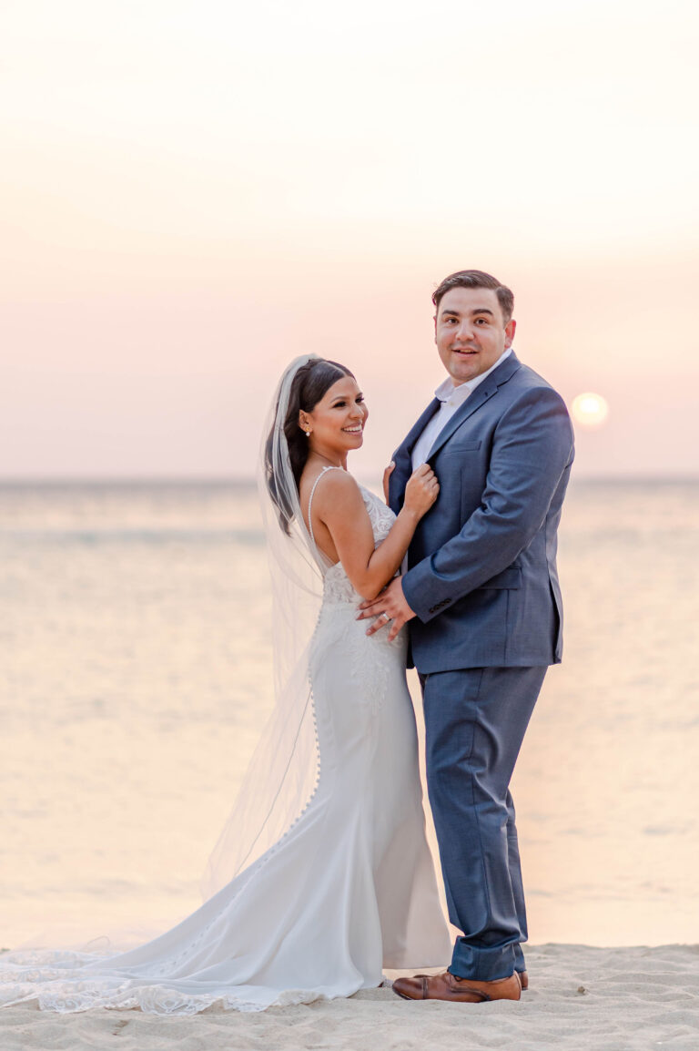 A newlywed couple stands on the sandy beach of Roatan, Honduras during sunset, with the ocean and setting sun in the background highlighting their wedding celebration.