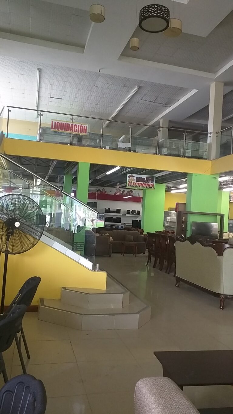 An indoor showroom in Roatan, Honduras features electronic appliances, furniture, and seating areas with bright green and yellow walls, and signs indicating liquidation and electronics categories.