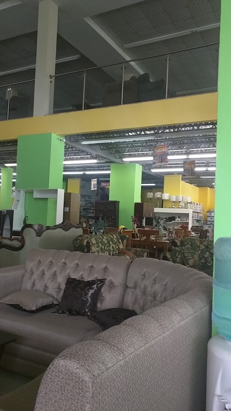 A furniture store in Roatan, Honduras features a variety of sofas and chairs with colorful cushions, displayed on a spacious floor with green and yellow pillars supporting an upper level, and retail sections visible in the background.