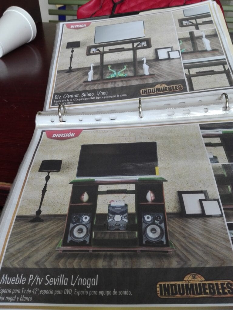 A black television stand with multiple shelves holding a flat-screen TV, speakers, and decorative swan sculptures, set against a beige wall and wooden flooring, displayed on a furniture catalog page for Roatan, Honduras.