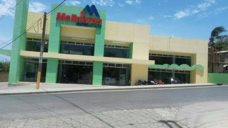 The image shows a colorful two-story building in Roatan, Honduras, with a sign reading "Molineros Supermercado," indicating a supermarket, featuring green and yellow exterior walls, large glass windows, and a green awning on the right side.