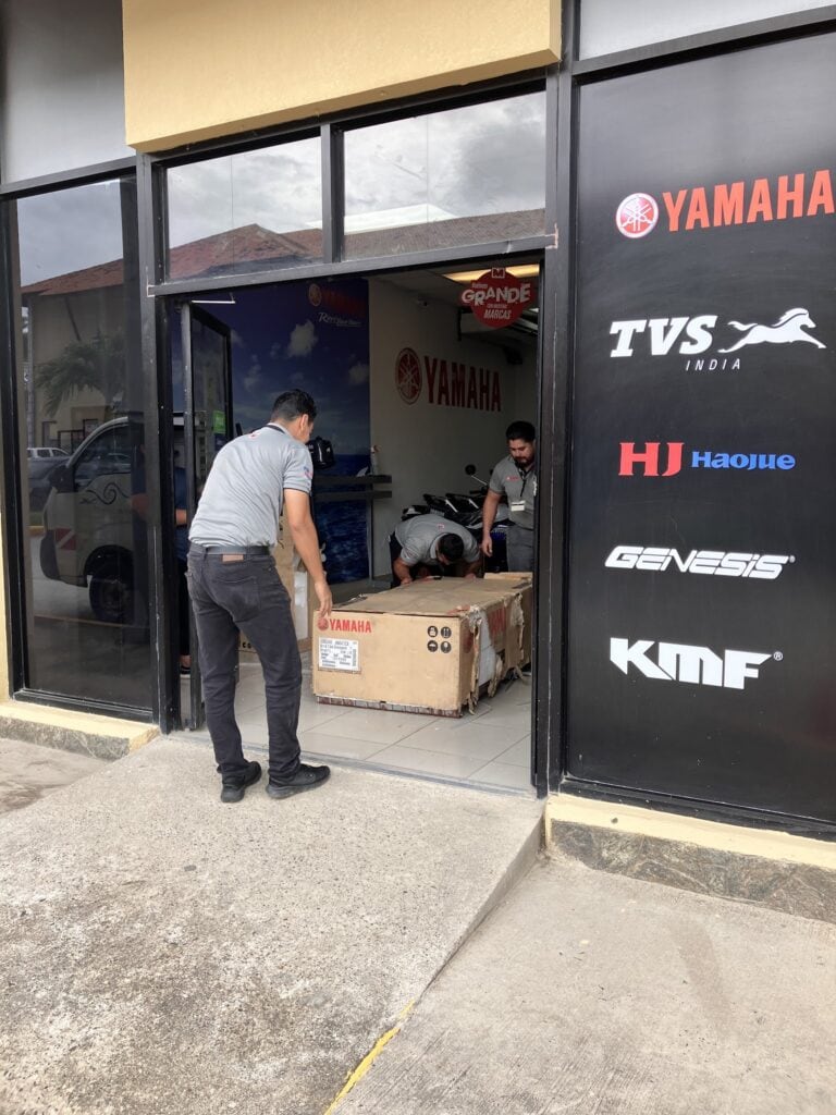 Two men are working inside a Yamaha motorcycle dealership in Roatan, Honduras, assembling or inspecting a Yamaha motorcycle box near the entrance, with a large Yamaha sign and other brand logos on the exterior wall.
