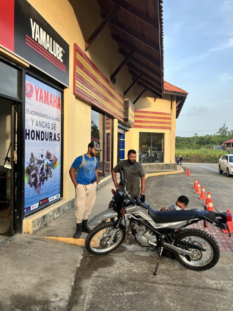 Three men are seen standing outside a Yamaha motorcycle dealership in Roatan, Honduras, with one man wearing a mask sitting near a Yamaha motorcycle while the other two stand nearby, against the backdrop of a yellow building with red and yellow decorative accents and a clear sky.