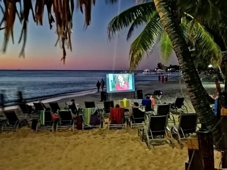 People sitting in chairs on a sandy beach in Roatan, Honduras, watch outdoor movies on a large screen at sunset, with palm trees and boats visible in the background.