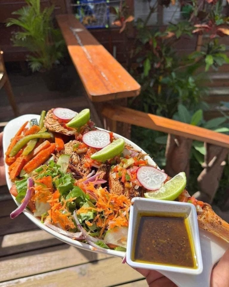 A plate of grilled fish topped with fresh vegetables, radish slices, lime wedges, and a side of sauce is held outdoors on a wooden deck with lush greenery in the background in Roatan, Honduras.