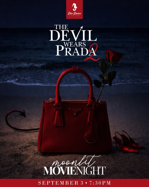 A red Prada handbag, a single red rose, and red high heels are arranged on a beach in Roatan, Honduras, with the ocean waves in the background, promoting the movie 'The Devil Wears Prada 2' at an event in September.