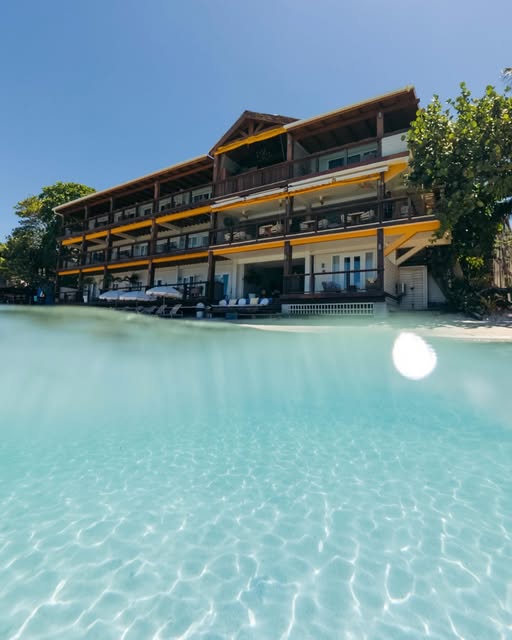 A beachfront hotel or resort in Roatan, Honduras, with multiple levels, balconies, and yellow accents, situated along clear turquoise waters under a bright blue sky with surrounding green trees.