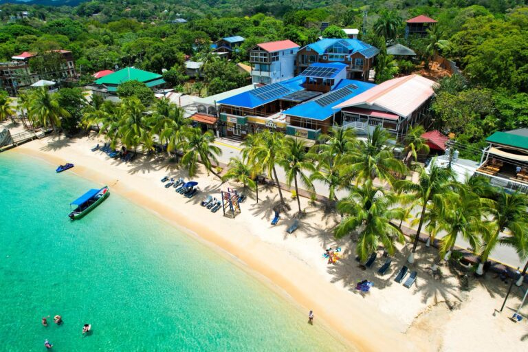 Aerial view of a beachfront resort in Roatan, Honduras, featuring colorful buildings with solar panels on the roofs, surrounded by lush green trees, with a sandy beach and clear turquoise waters where people are swimming and relaxing.
