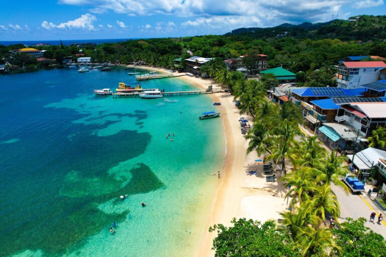 Aerial view of a beachfront area in Roatan, Honduras, featuring a clear turquoise bay with boats docked at a wooden pier, surrounded by lush green trees, colorful houses, and buildings with solar panels on their rooftops along a sandy shoreline.
