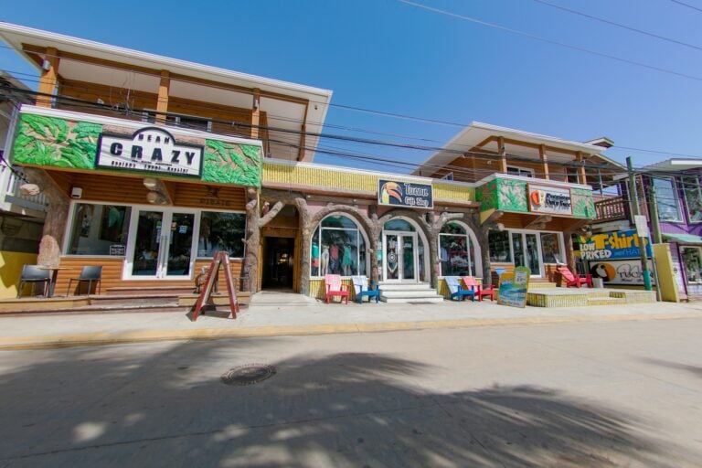 A colorful two-story business building in Roatan, Honduras, features a vibrant sign for Bean Crazy, a gift shop, and a restaurant, with outdoor seating, tropical decor, and signage advertising T-shirts and souvenirs.