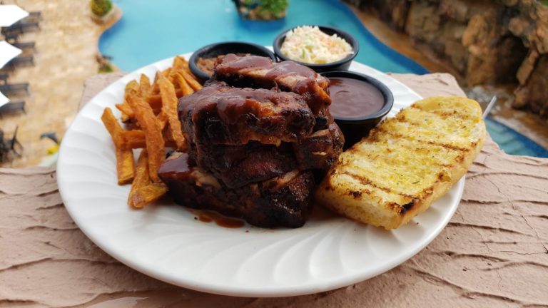 A plate of traditional Honduran barbecue with grilled bread, French fries, coleslaw, and barbecue sauces is shown outdoors in Roatan, Honduras, with a swimming pool and rocky surroundings in the background.