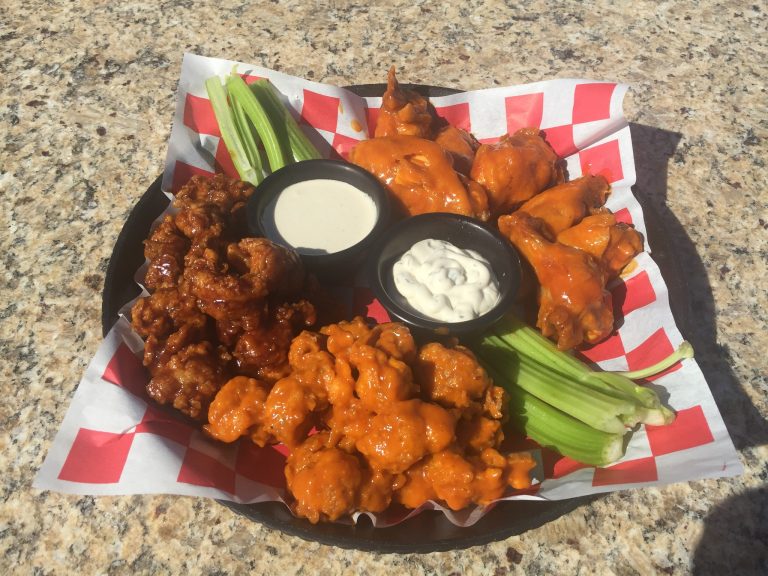 A platter of spicy chicken wings with celery sticks, dipping sauces, and a creamy dressing is displayed on a checkered paper at a restaurant in Roatan, Honduras.