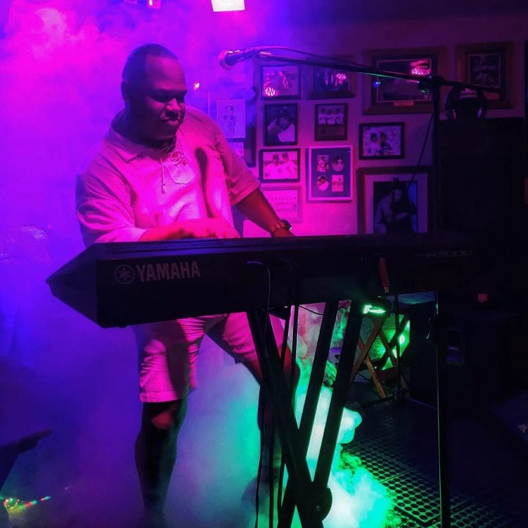 A musician performs on a Yamaha keyboard in a dimly lit, colorful bar or lounge in Roatan, Honduras, with neon lights illuminating the scene and framed photos on the wall behind him.