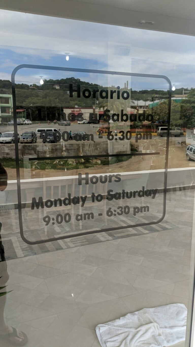 A business hours sign displayed on a glass door shows that the establishment in Roatan, Honduras, is open Monday to Saturday from 9:00 am to 6:30 pm, with the background reflecting a parking lot, buildings, and green hills under a partly cloudy sky.