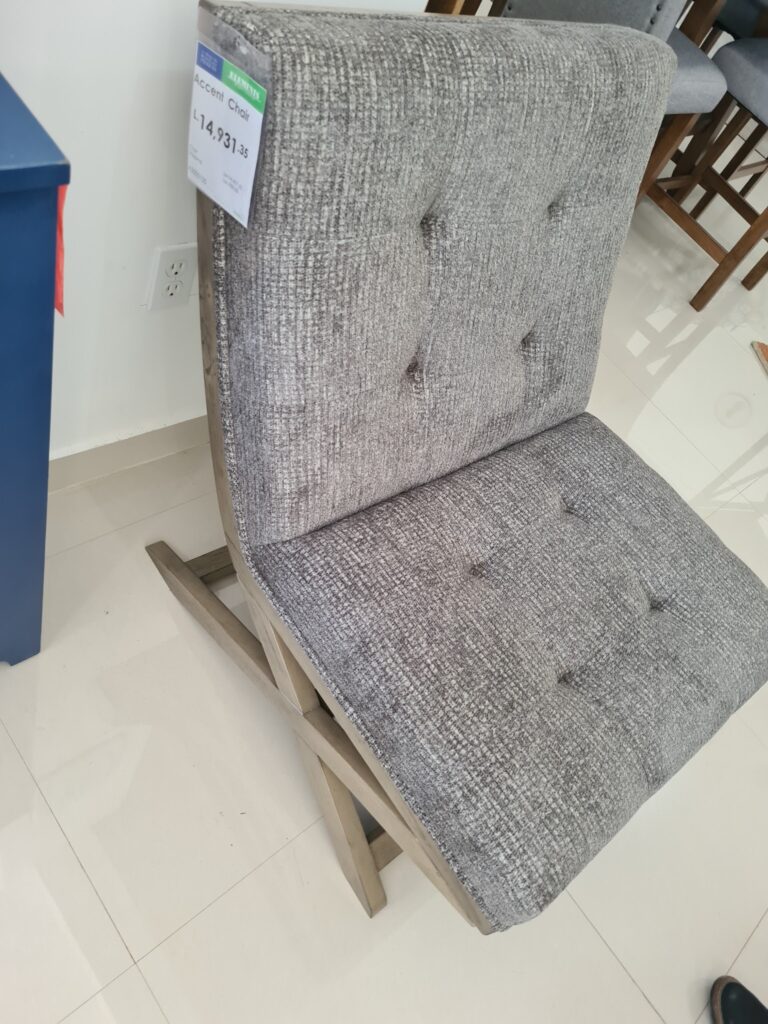 A gray textured accent chair with a wooden frame is displayed indoors at a furniture store in Roatan, Honduras, with a price tag attached.