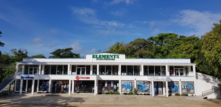 A two-story white retail building in Roatan, Honduras, displays signage for "Elements" furniture store on the top, with storefronts for clothing and accessories, including a "Pacer" store and a mural of vintage cars on the lower level, surrounded by lush green trees and a bright blue sky.