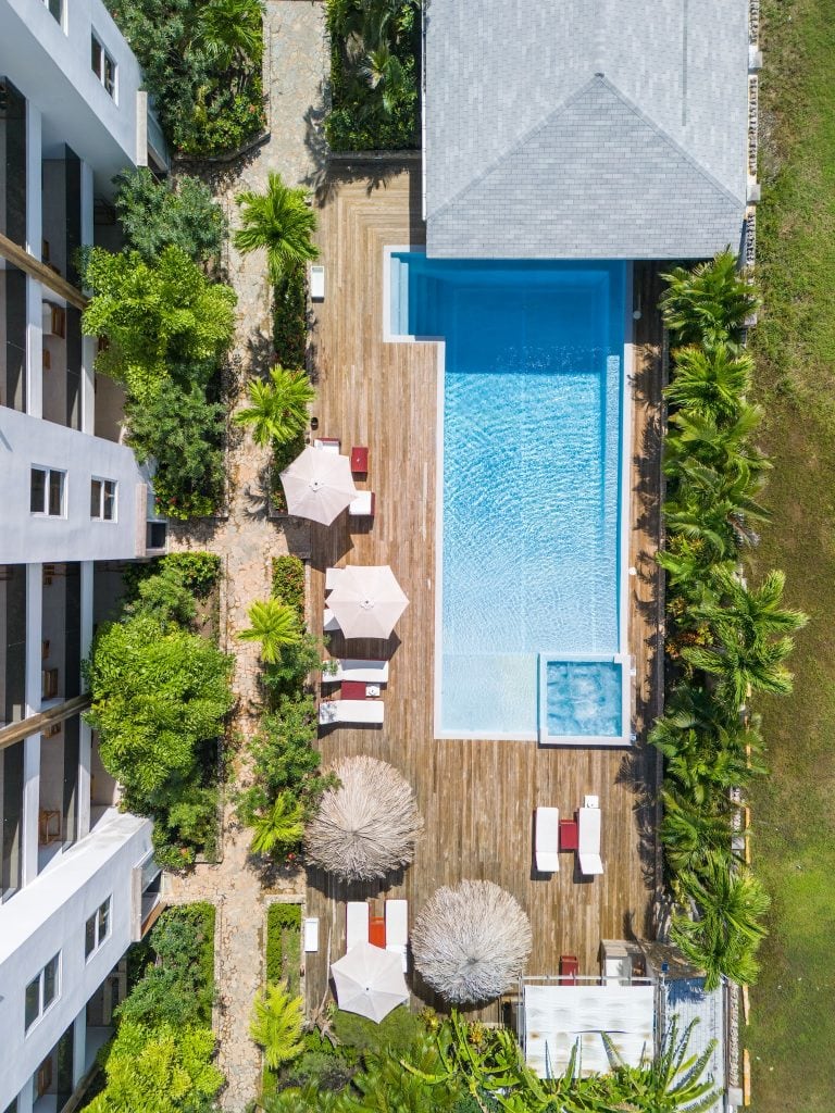 An aerial view of a swimming pool surrounded by lush tropical plants, white umbrellas, and wooden decking, with a modern hotel building on the left and green grassy areas in Roatan, Honduras.