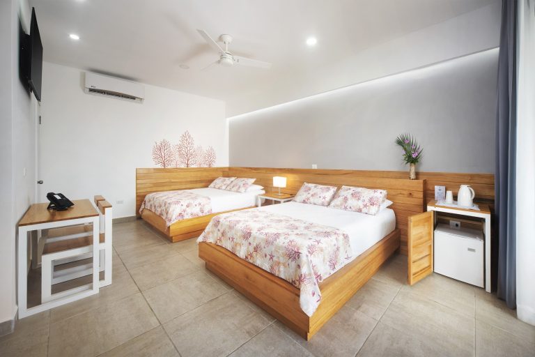 A modern hotel room in Roatan, Honduras features two single beds with sea-themed pink and white bedding, a wooden headboard, a small bedside table with a lamp, a wall-mounted TV, a work desk with a telephone, a vase with flowers, and a ceiling fan with an air conditioning unit.