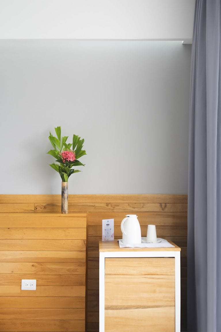 A white wall with a potted pink flower and green leaves on a wooden ledge, a small white table holding a white pitcher and cups, in a modern interior space in Roatan, Honduras.