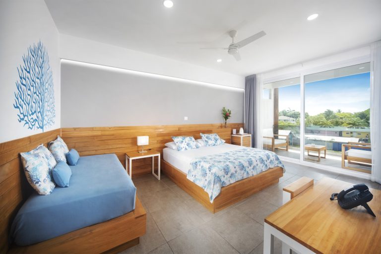 A modern hotel room on Roatan, Honduras, features three beds with blue and floral bedding, a large window with a view of tropical trees, and minimalist decor including a blue coral wall art and wooden accents.