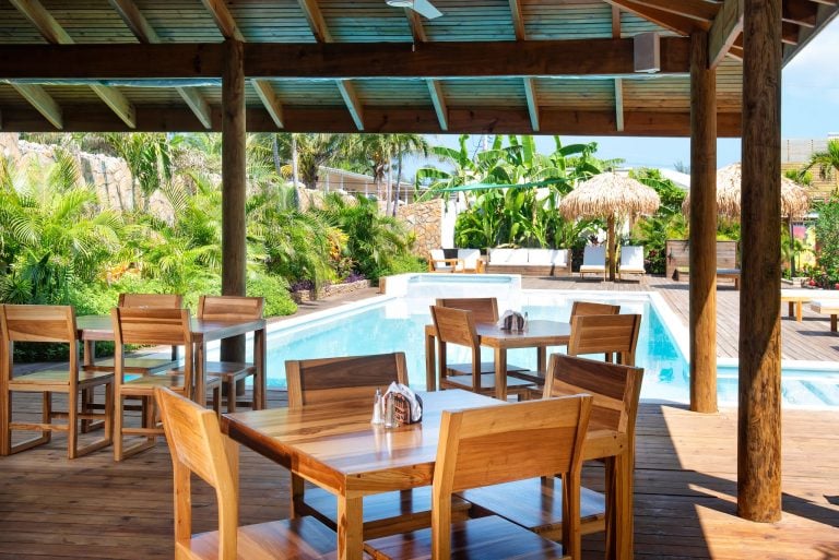 A shaded outdoor dining area with wooden tables and chairs overlooks a swimming pool, surrounded by lush tropical plants and palm trees on Roatan, Honduras.