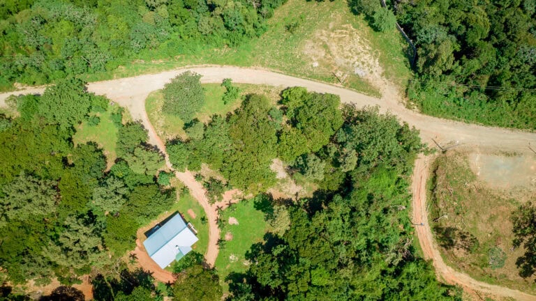 Aerial view of a small white house with a blue roof on grassy land surrounded by dense green trees on Roatan, Honduras, with a dirt road winding through the property.