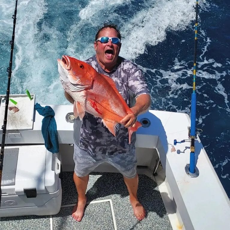 A man holding a large red fish on a boat in clear blue waters off Roatan, Honduras, with fishing rods and equipment visible on the boat deck.