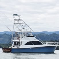 A large white and blue fishing or tour boat is anchored on the water near Roatan, Honduras, with lush green mountains and a cloudy sky in the background.