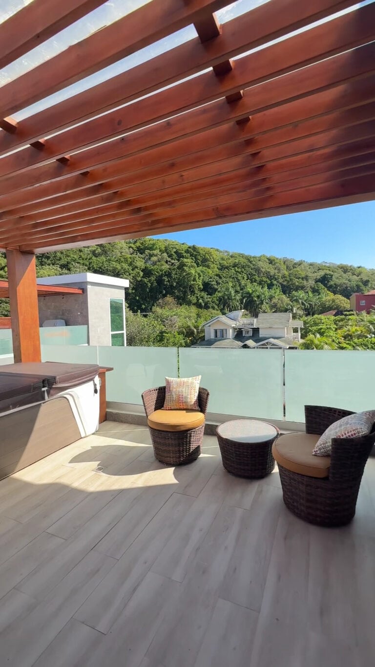 A rooftop patio in Roatan, Honduras, features wicker chairs with cushions, a hot tub, and a view of lush green hills and residential houses beneath a wooden pergola.
