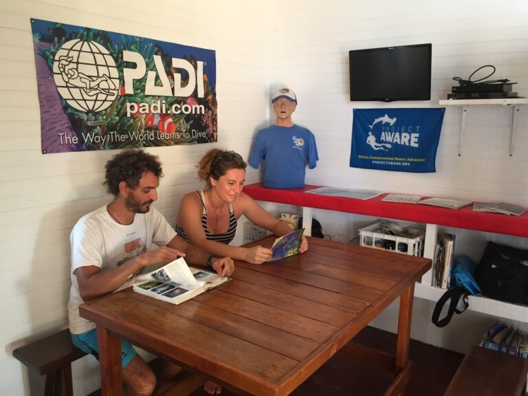 Two people sit at a wooden table inside a building on Roatan, Honduras, reading brochures or maps, with PADI diving banners and a television mounted on the white-paneled wall behind them.
