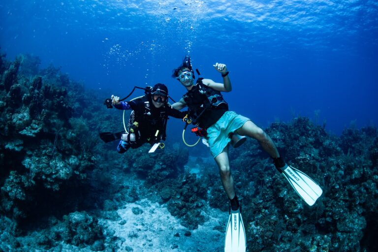 Two scuba divers pose underwater over vibrant coral reefs in Roatan, Bay Islands, Honduras, with one giving a thumbs-up and the other making an OK sign, during a diving adventure in the Caribbean Sea.