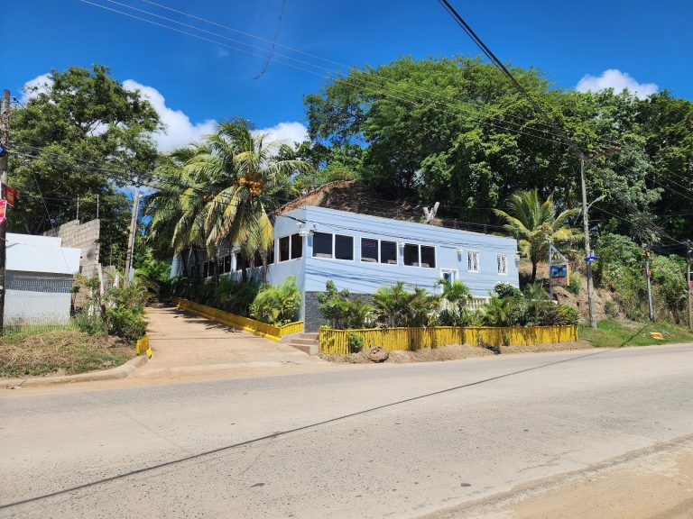 A white, two-story building with large windows and an "open" sign, surrounded by lush green trees and palm trees on a hillside in Roatan, Honduras, with electrical wires overhead and a paved sidewalk in front.