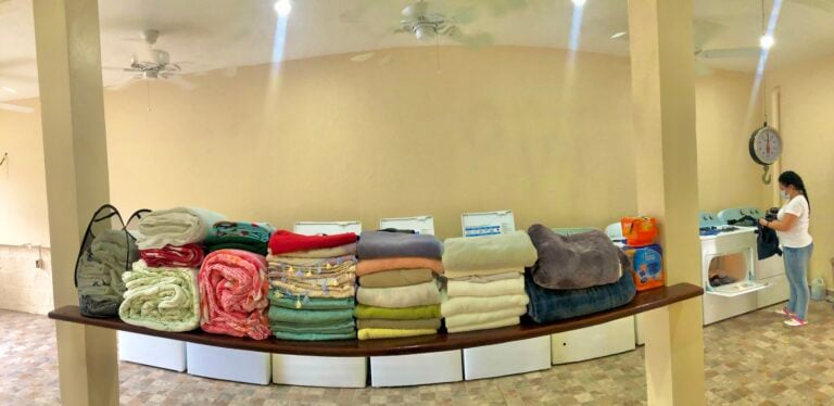 Laundry room in Roatan, Honduras with stacked towels and blankets, washing machines, and a woman handling laundry while wearing a face mask.
