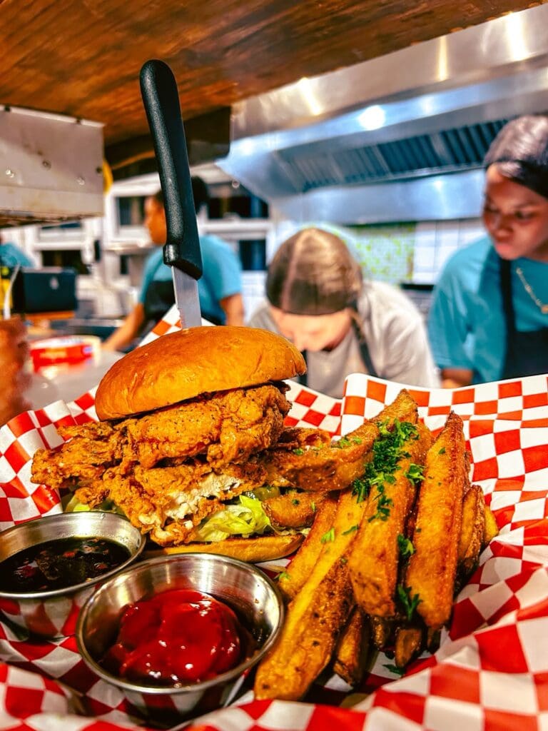 Fried chicken sandwich with crispy chicken filet, lettuce, and bun, served with seasoned fried plantains and dipping sauces at a restaurant in Roatan, Honduras.