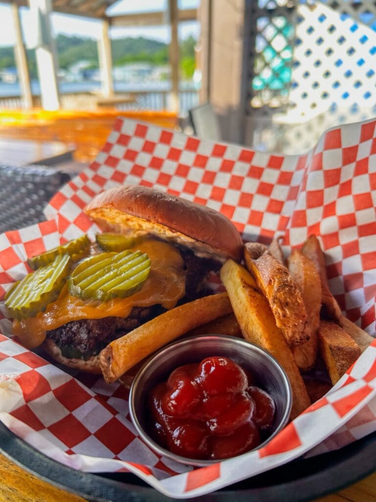 Close-up of a delicious cheeseburger with pickles, melted cheese, lettuce, and a toasted bun, served with thick-cut fries and ketchup on a metal tray lined with red and white checkered paper in an outdoor seating area on Roatan, Bay Islands, Honduras.