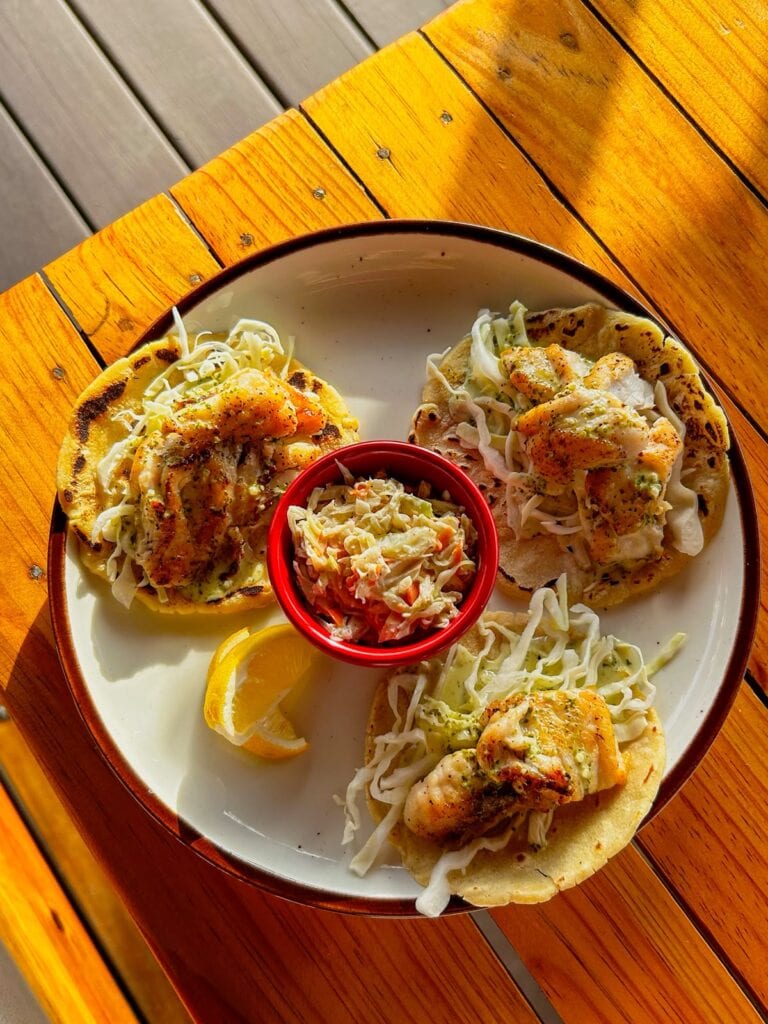 Three fish tacos topped with shredded cabbage and sauce served on a white plate with a lemon wedge and a side of coleslaw on a wooden table in Roatan, Honduras.